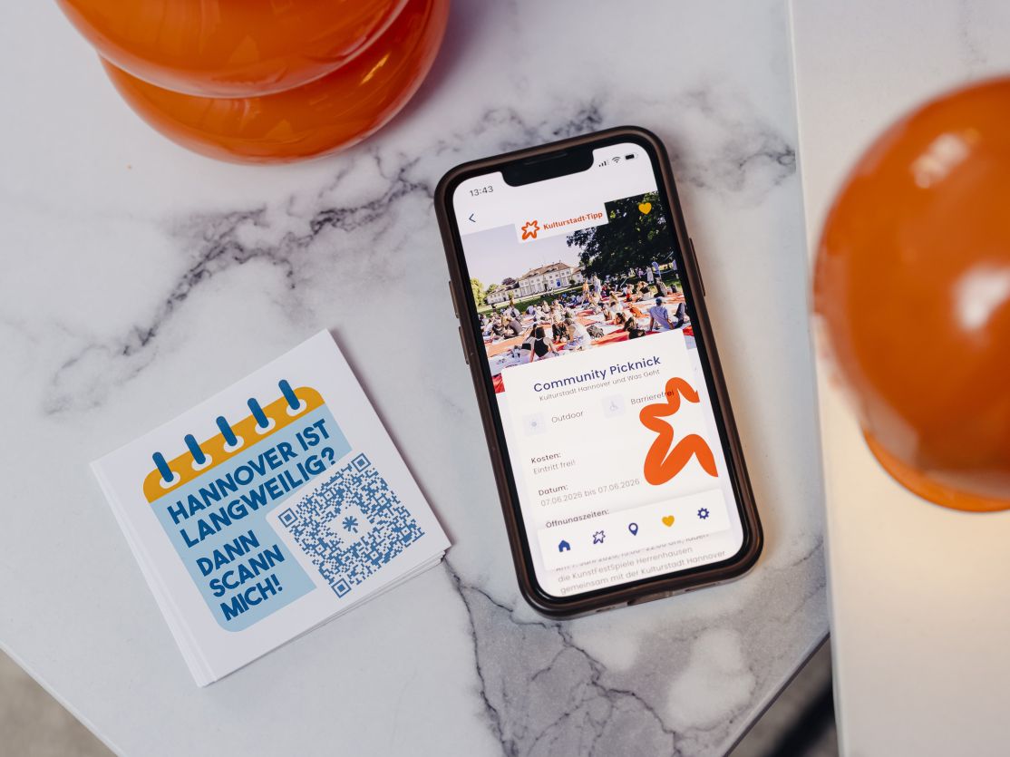 Smartphone zeigt App mit "Community Picknick", daneben Flyer mit QR-Code und Text "Hannover ist langweilig? Dann scann mich!"