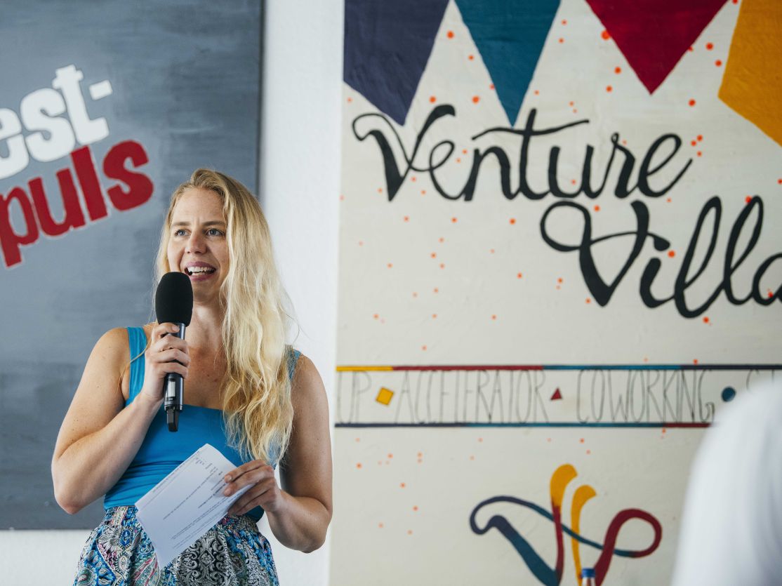 Blonde Frau mit Mikrofon spricht vor einem Venture Villa Schild, hält Papier in der Hand.