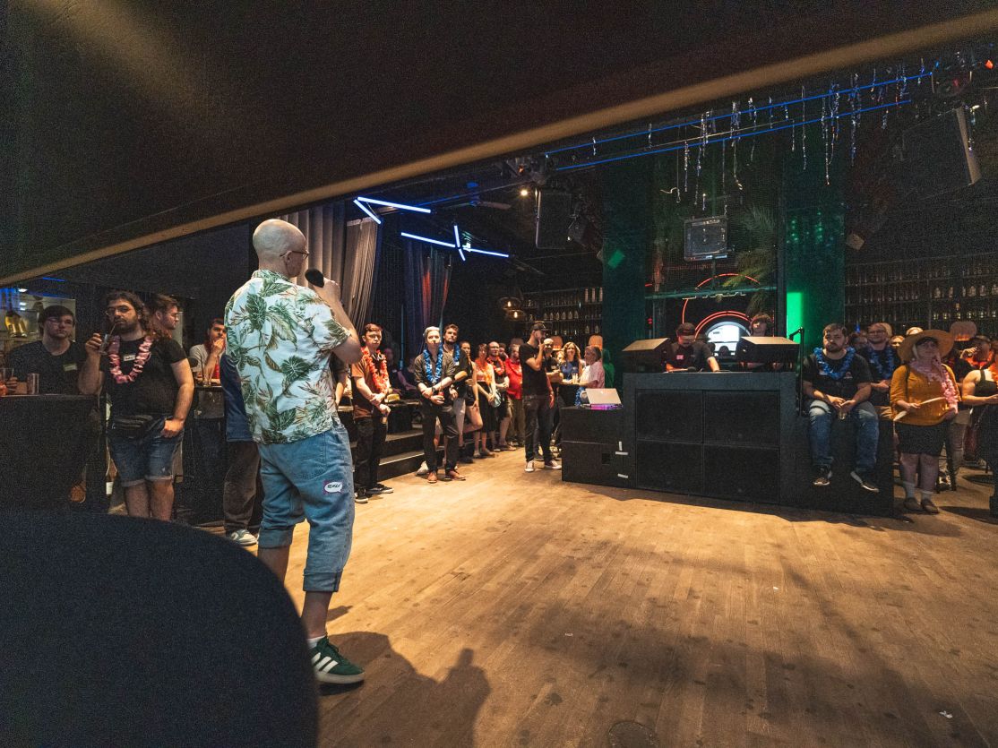 Person in Hawaiihemd steht auf Bühne vor Publikum in dunklem Club mit Holzboden und Neonbeleuchtung.