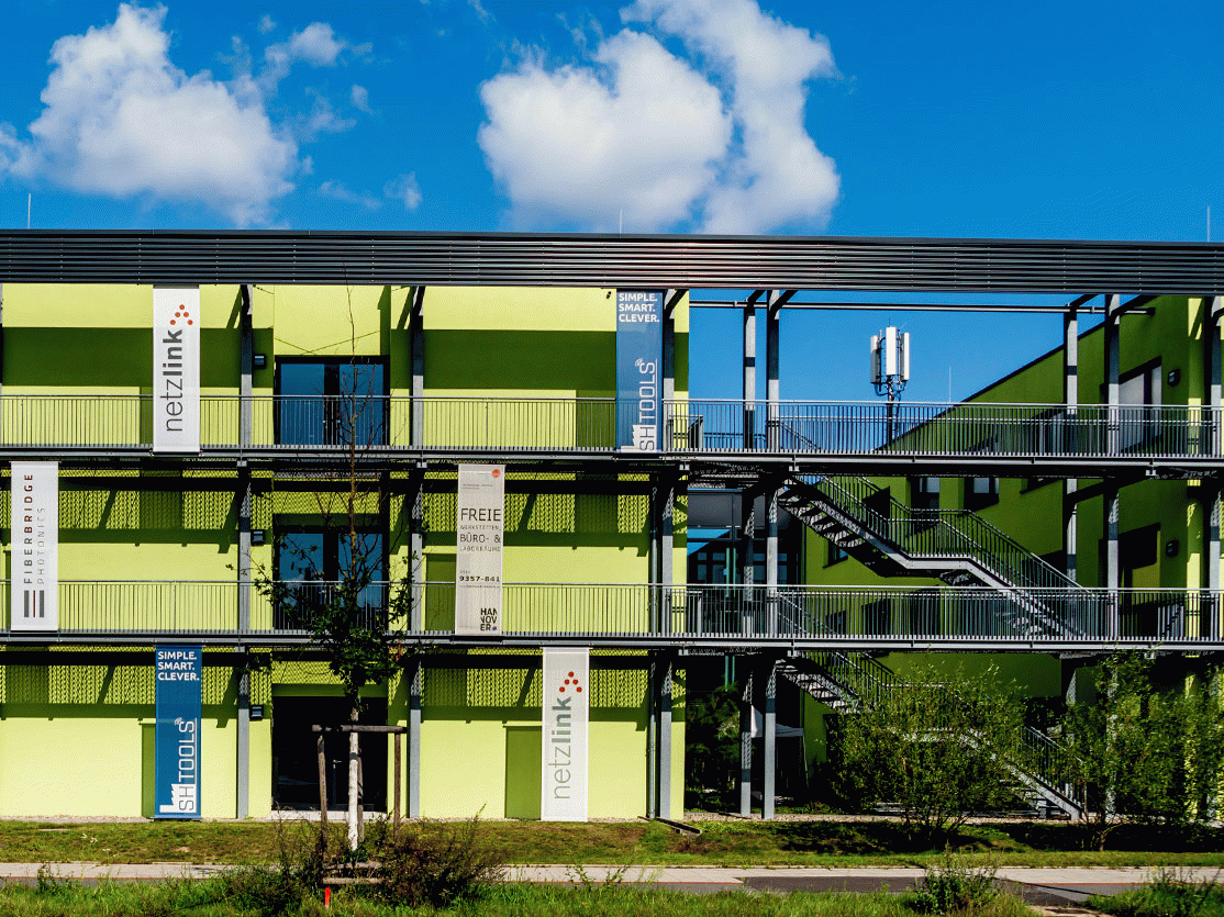 Grünes Bürogebäude mit mehreren Etagen, Balkonen und Firmenschildern „netzlink" und „ST tools", blauer Himmel.