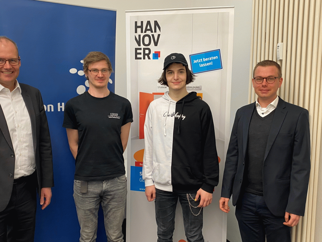 Vier Männer stehen vor einem Hannover-Banner. Zwei tragen Anzüge, zwei Jüngere sind casual gekleidet.