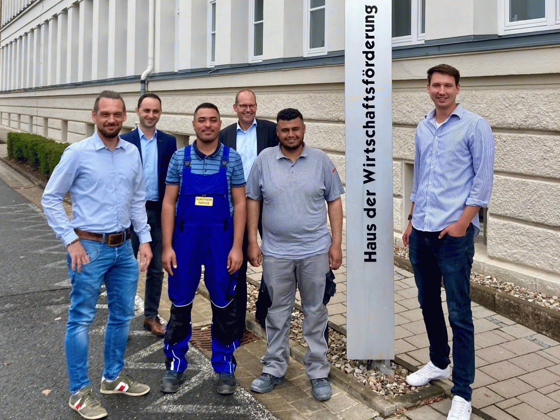 Sechs Männer stehen vor dem Schild „Haus der Wirtschaftsförderung". Zwei tragen Arbeitskleidung, die anderen Freizeitkleidung.