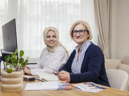 Zwei Frauen sitzen an einem Schreibtisch mit Computer, Dokumenten und Pflanze; eine trägt Hijab, die andere Brille.