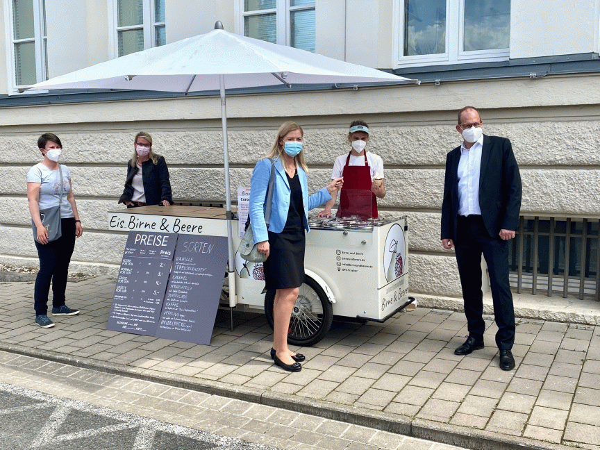 Fünf Personen mit Masken stehen um einen Eiswagen „Eis, Birne & Beere" mit weißem Sonnenschirm auf einem Gehweg.