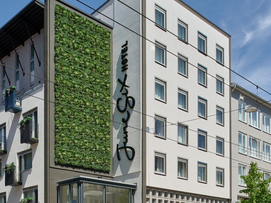 Modernes Stadtgebäude mit begrünter Fassadenwand, Glaseingang, Fahrrädern und Fußgängern auf dem Gehweg.