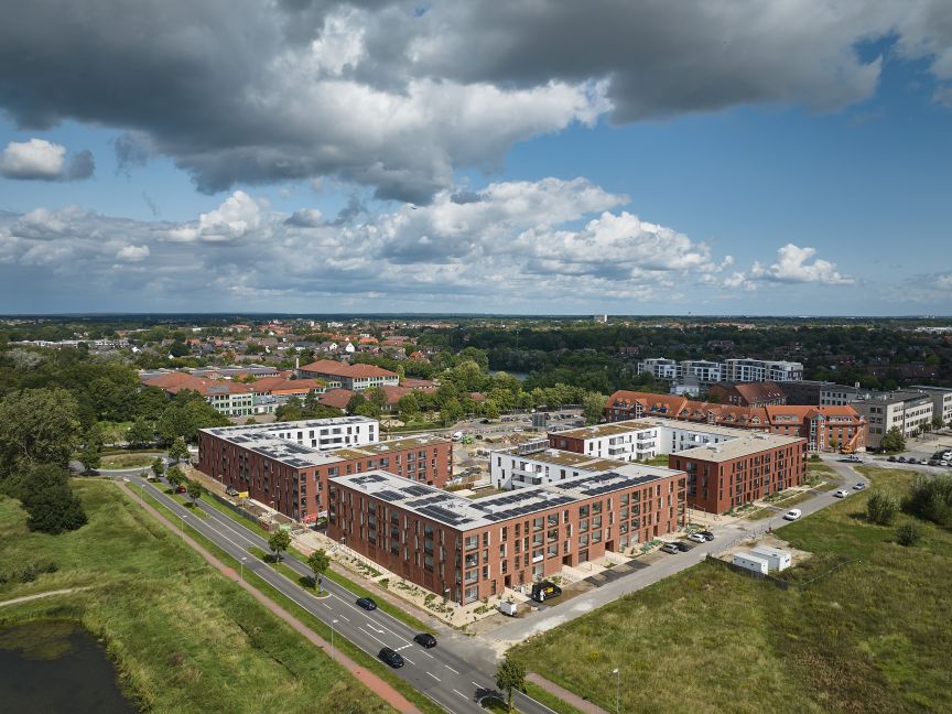 Luftaufnahme eines Stadtviertels mit roten Backsteingebäuden, Solaranlagen auf Dächern, Straßen und grüner Umgebung.