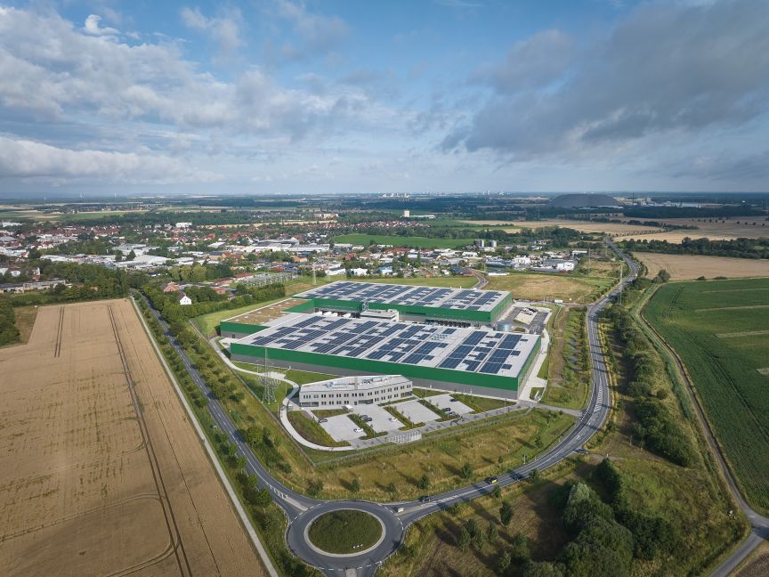 Luftaufnahme eines großen Industriegebäudes mit Solarpanelen auf dem Dach, umgeben von Feldern und einer Kleinstadt.