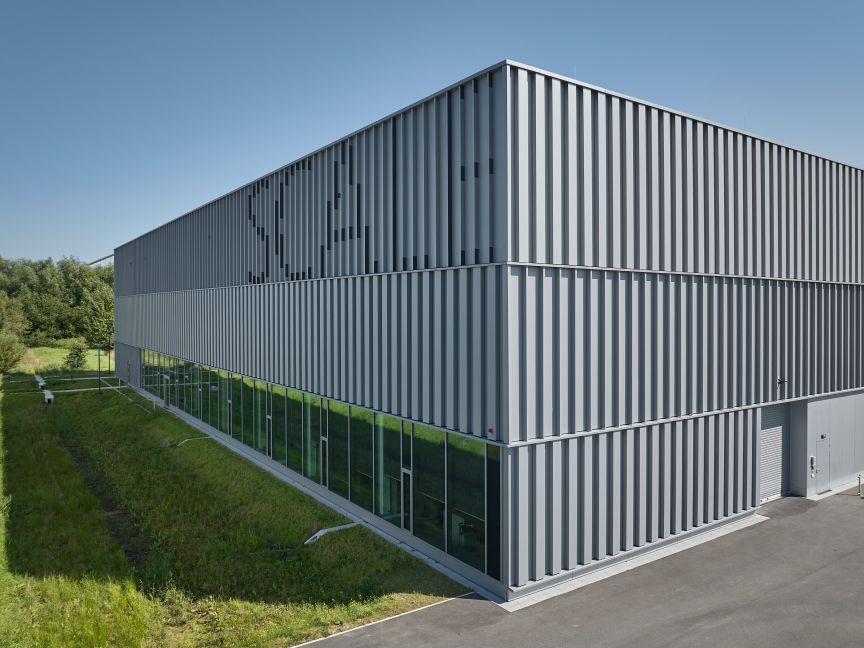 Modernes Industriegebäude mit grauer Metallfassade, vertikalen Lamellen und Glasfront im Erdgeschoss, umgeben von Grünfläche.