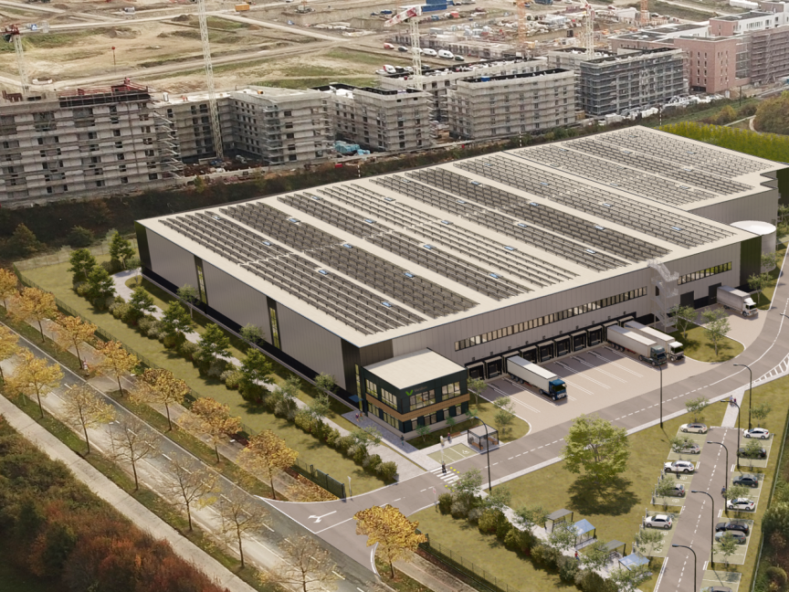Luftbild eines großen Lagergebäudes mit Solardach, Laderampen, LKW und umliegenden Wohngebäuden im Bau.
