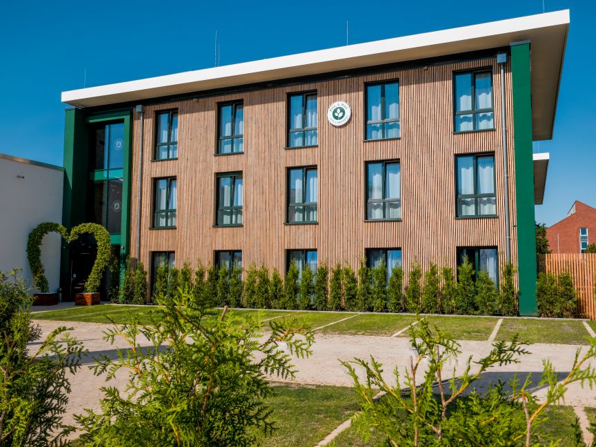 Modernes dreigeschossiges Gebäude mit Holzfassade und grünen Akzenten, umgeben von Büschen und Bäumen bei blauem Himmel.