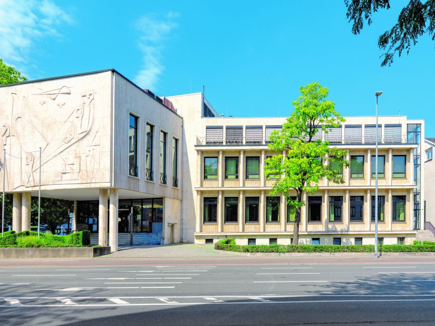 Modernes Verwaltungsgebäude mit Reliefschmuck, mehrstöckiger Fassade, Bäumen und breiter Straße bei blauem Himmel.