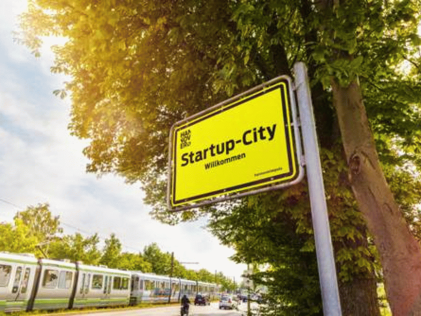 Gelbes Ortsschild „Startup-City – Willkommen" an einem Metallpfahl neben einem Baum, im Hintergrund Straßenbahn und Bäume.