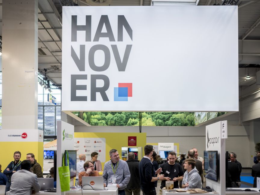 Messestand mit großem „HANNOVER"-Schriftzug, darunter Besucher an Tischen mit Laptops in einer Ausstellungshalle.