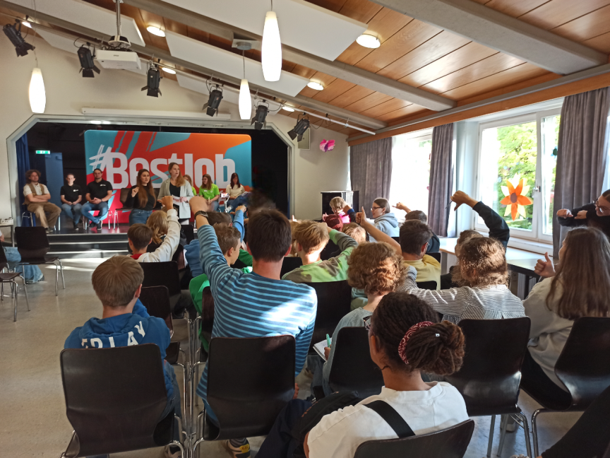 Kinder sitzen in einem Saal und strecken die Hand auf. Auf einer Bühne mit "#Bestjob"-Banner sitzen mehrere Erwachsene.