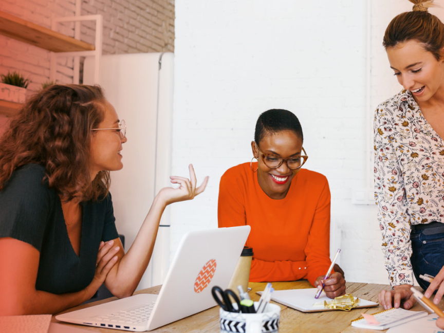 Drei Frauen arbeiten gemeinsam an einem Tisch mit Laptop und Notizen in einem Büro.