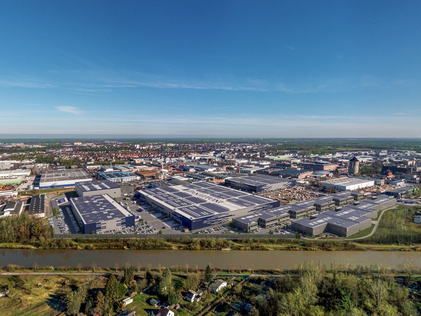 Luftaufnahme eines Industriegebiets mit großen Lagerhallen, Parkplätzen und einem Fluss im Vordergrund.