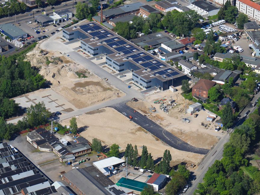 Luftaufnahme eines Industriegebiets mit Baustelle, Sandgelände, Flachdachgebäuden mit Solaranlagen und umliegender Bebauung.