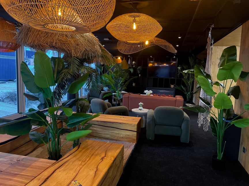 Lounge mit Rattanlampen, grünen Sesseln, Holztischen, Topfpflanzen und einem Flachbildschirm an dunkler Wand.
