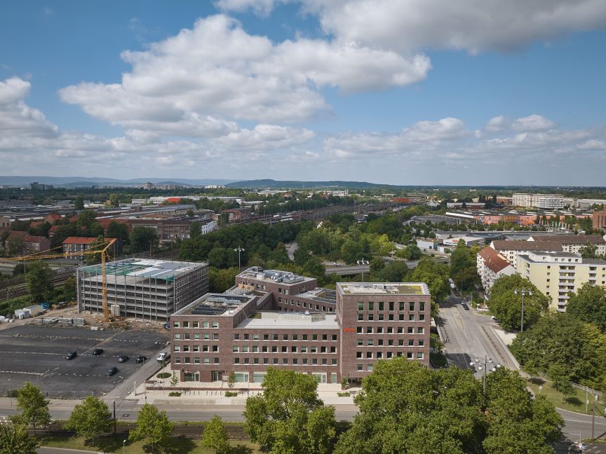 Luftaufnahme einer Stadt mit Bürogebäuden, Baustelle mit Kran, Straßen, Bäumen und Hügeln im Hintergrund bei bewölktem Himmel.