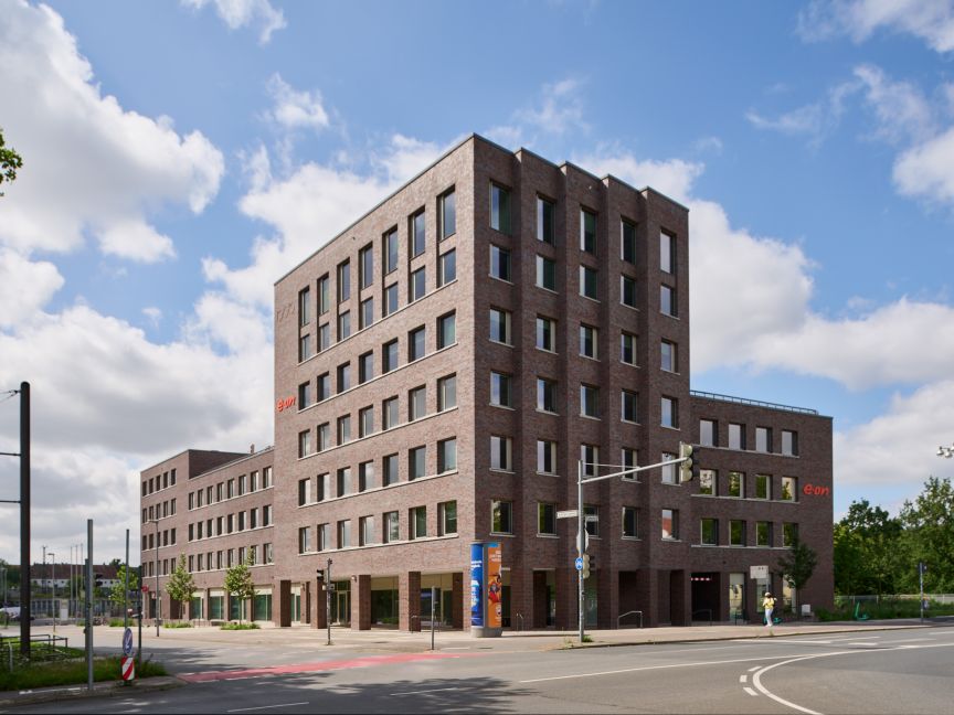 Modernes mehrstöckiges Backsteingebäude an einer Straßenkreuzung mit Ampel, blauer Himmel mit Wolken.