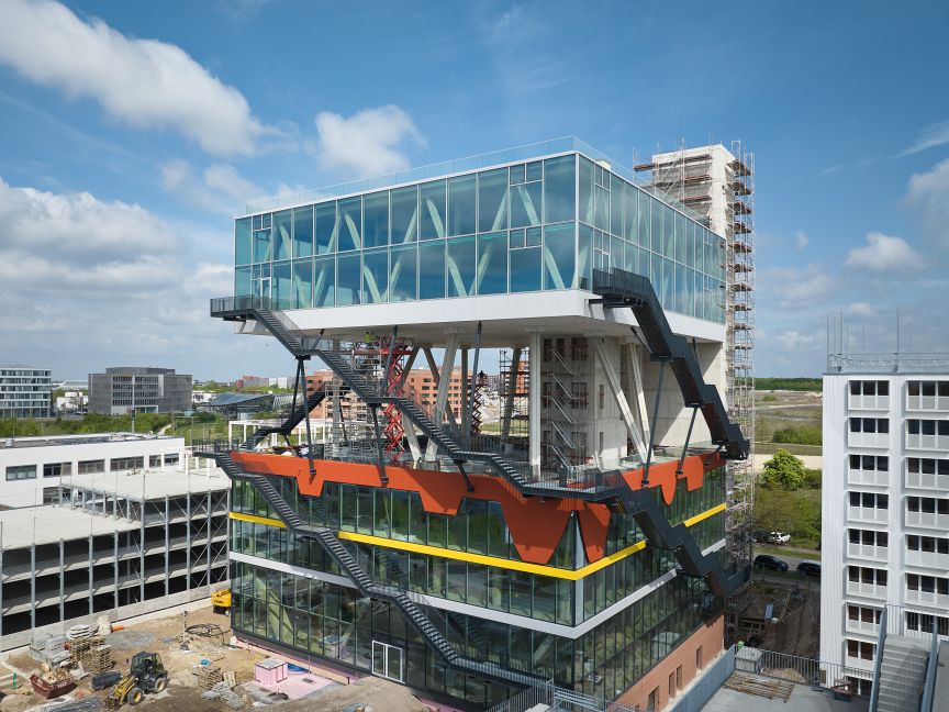 Modernes Glasgebäude mit orangefarbenen Stahlträgern und außenliegenden Treppen, umgeben von Baustellen und Stadtgebäuden.
