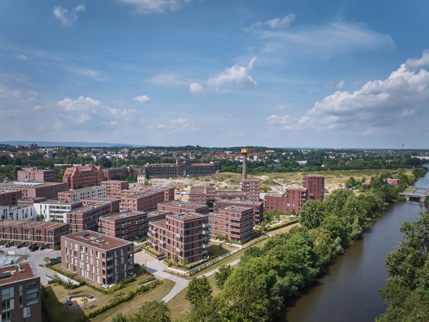 Luftaufnahme einer Stadt mit roten Backsteingebäuden, einem Fluss, Industrieschornstein und grünen Bäumen unter blauem Himmel.