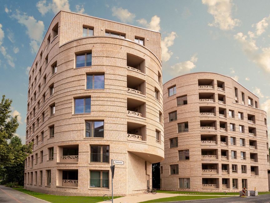 Modernes mehrstöckiges Wohngebäude aus hellem Backstein mit geschwungener Fassade, Balkonen und großen Fenstern, blauer Himmel.