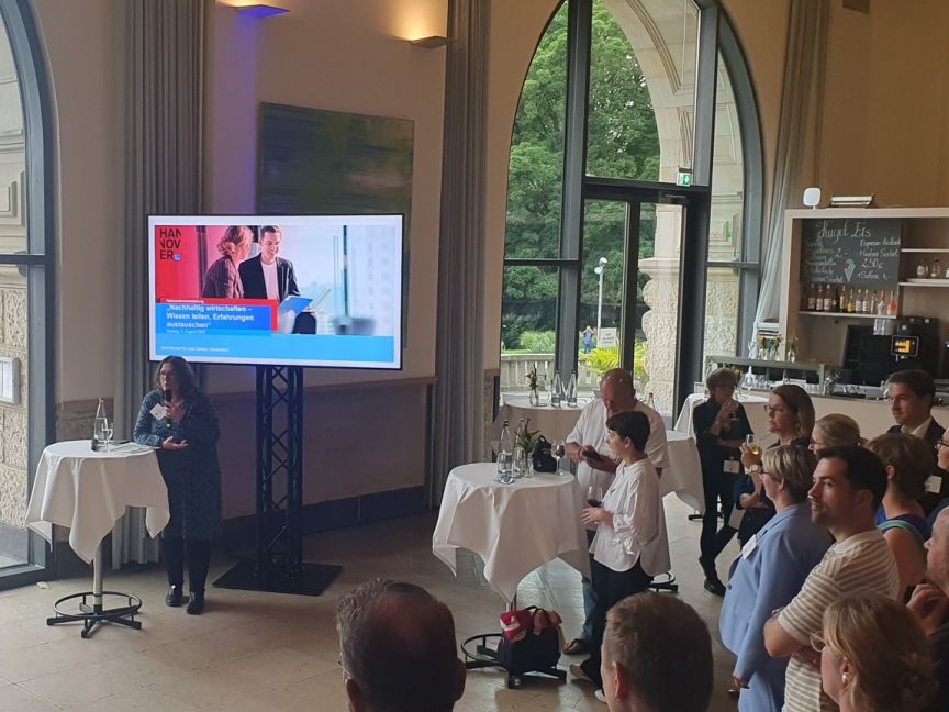 Frau spricht am Podium vor Publikum in elegantem Saal. TV-Bildschirm zeigt Präsentation mit Logo „Hannover".