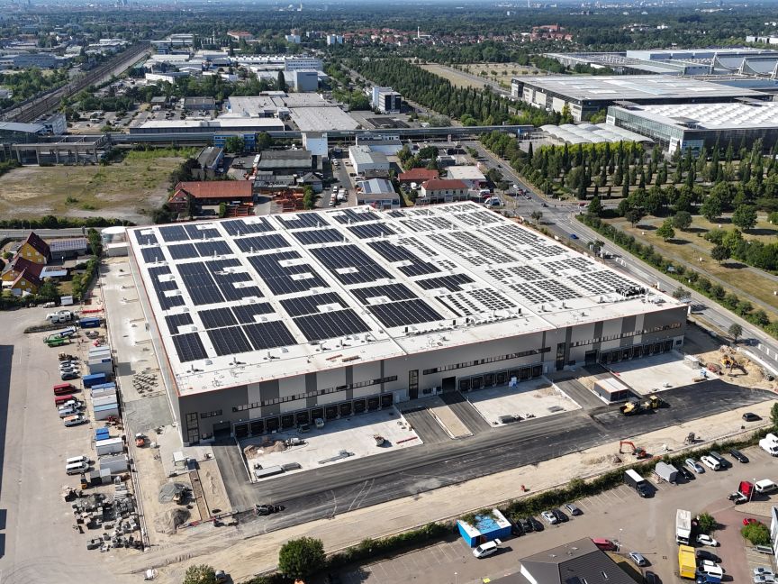 Luftaufnahme eines großen Industriegebäudes mit Solaranlage auf dem Flachdach, umgeben von Industriegebiet und Grünflächen.