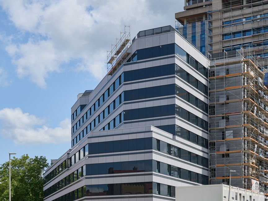 Modernes Bürogebäude mit blau-weißer Fassade neben einem Hochhaus mit Baugerüst, davor Straße und Zaun.