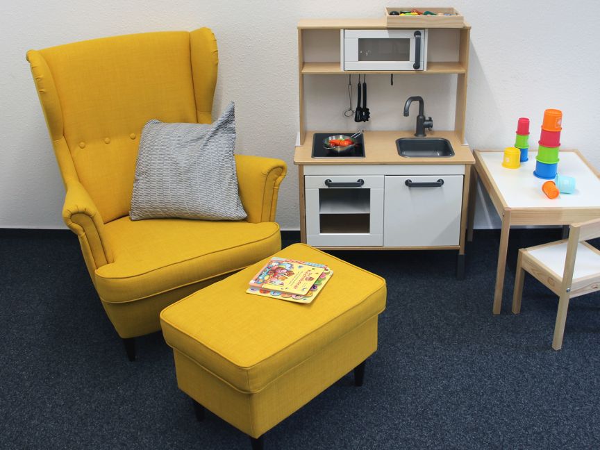 Gelber Sessel mit Hocker und grauem Kissen, daneben Spielküche und Kindertisch mit bunten Stapelbechern.