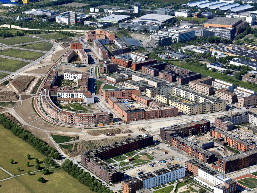 Luftaufnahme einer Stadtentwicklung mit mehrstöckigen Wohngebäuden, Baustellen, Grünflächen und Industriegebäuden im Hintergrund.