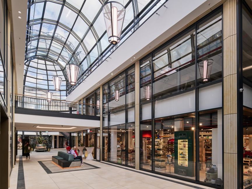 Helles Einkaufszentrum mit Glasdach, eleganten Hängelampen, zwei Etagen, Geschäften hinter Glasfronten und Sitzmöbeln im Gang.