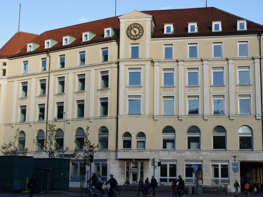 Mehrstöckiges Gebäude mit cremefarbener Fassade, rotem Dach und Uhr, belebt von Fußgängern an einer Straßenecke.