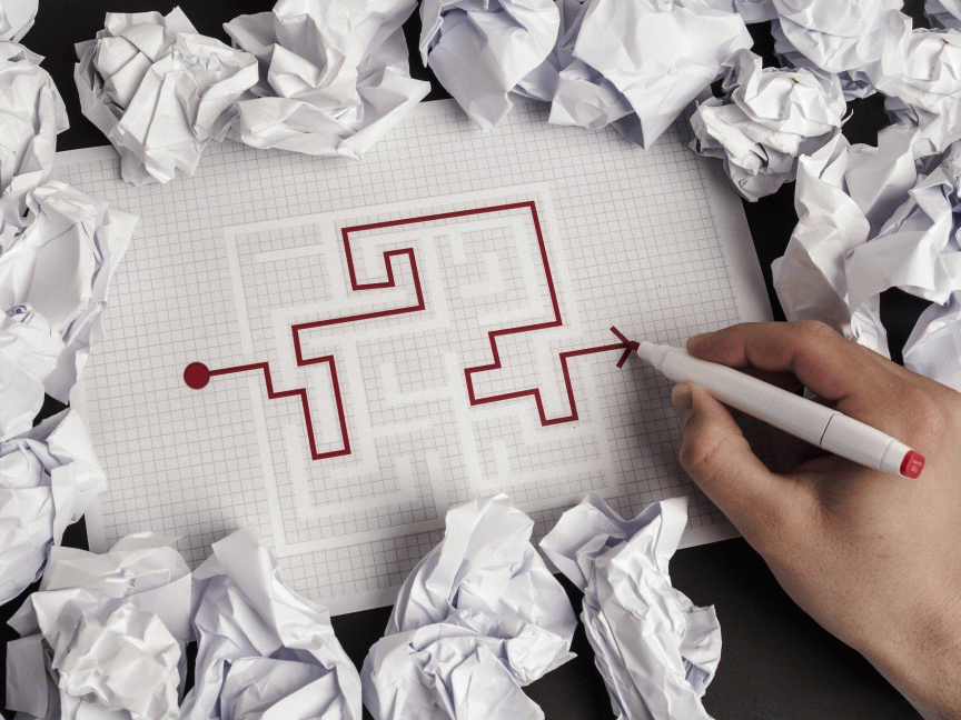Hand zeichnet mit rotem Marker einen Lösungsweg durch ein Labyrinth auf kariertem Papier, umgeben von zerknüllten Blättern.