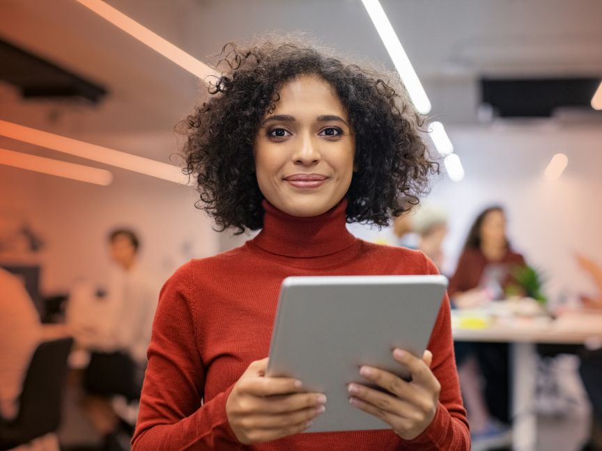 Frau mit lockigem Haar und rotem Rollkragenpullover hält Tablet in einem modernen Büro.