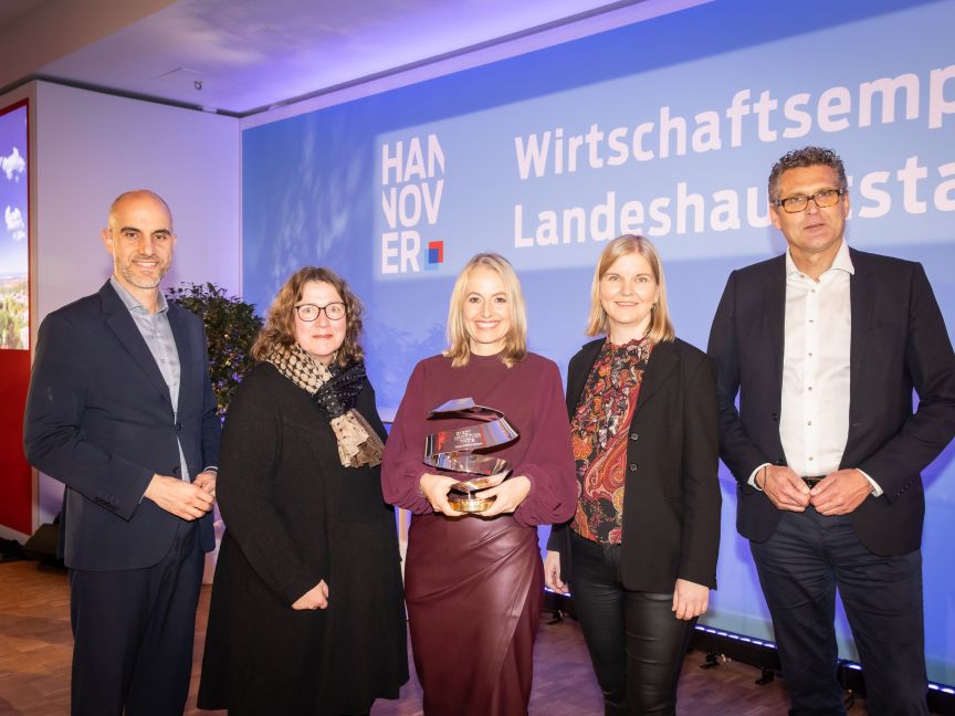 Fünf Personen stehen vor einem Hannover-Banner. Eine Frau in der Mitte hält eine Auszeichnung in den Händen.