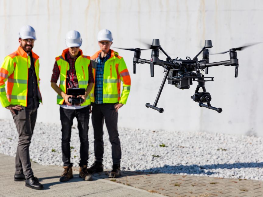 Drei Personen in Warnwesten und Helmen steuern eine Drohne mit Kamera auf einer Baustelle.