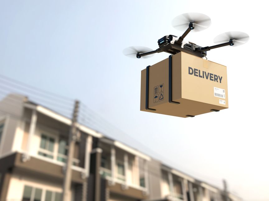 Drohne fliegt mit einem Karton mit Aufschrift „DELIVERY" über Wohnhäuser vor hellem Himmel.