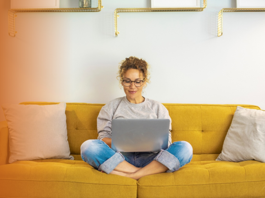 Frau mit Brille sitzt im Schneidersitz auf gelbem Sofa und arbeitet an einem Laptop.