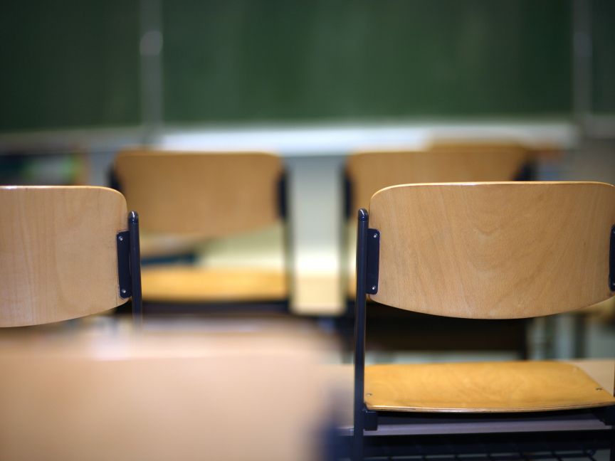 Leere Holzstühle in einem Klassenzimmer mit grüner Tafel im Hintergrund.
