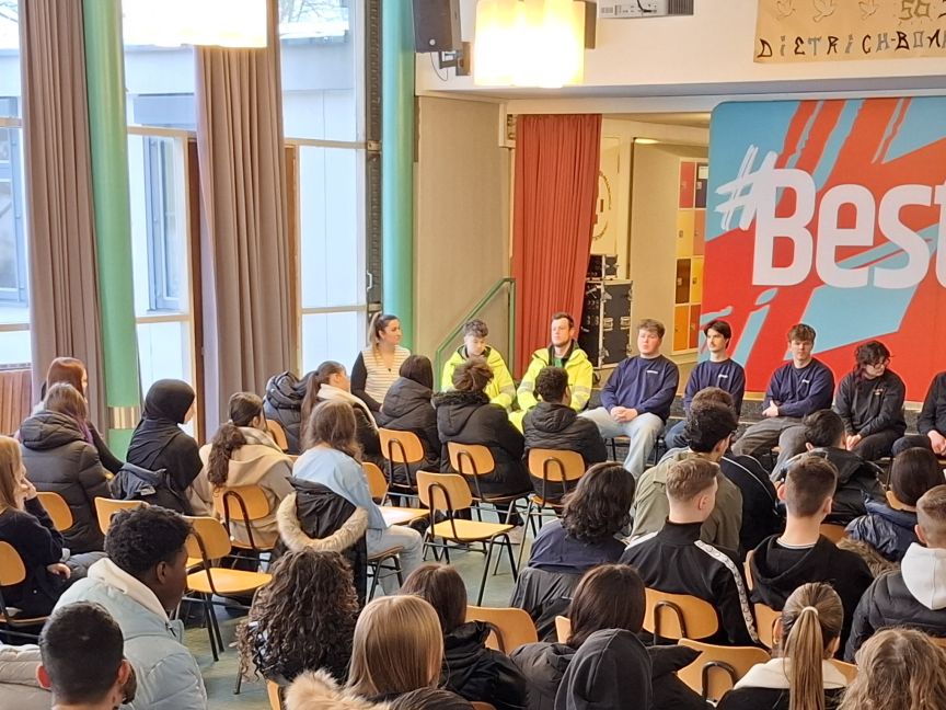 Schüler sitzen in Stuhlreihen vor einem Panel mit Erwachsenen in einer Aula. Banner mit "#BestJob Ever" im Hintergrund.
