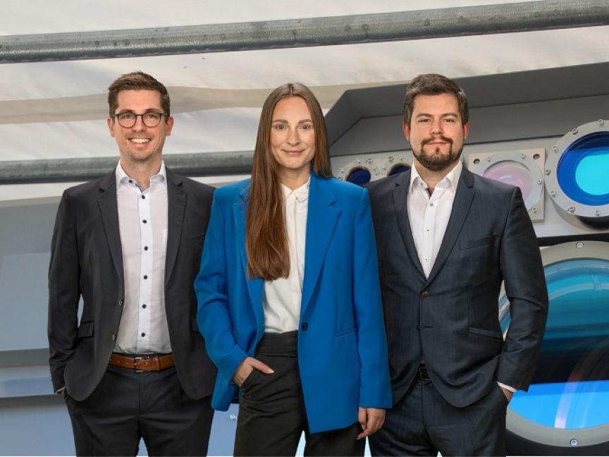 Drei Personen in Businesskleidung stehen lächelnd vor einem technischen Gerät mit blauer Optik.