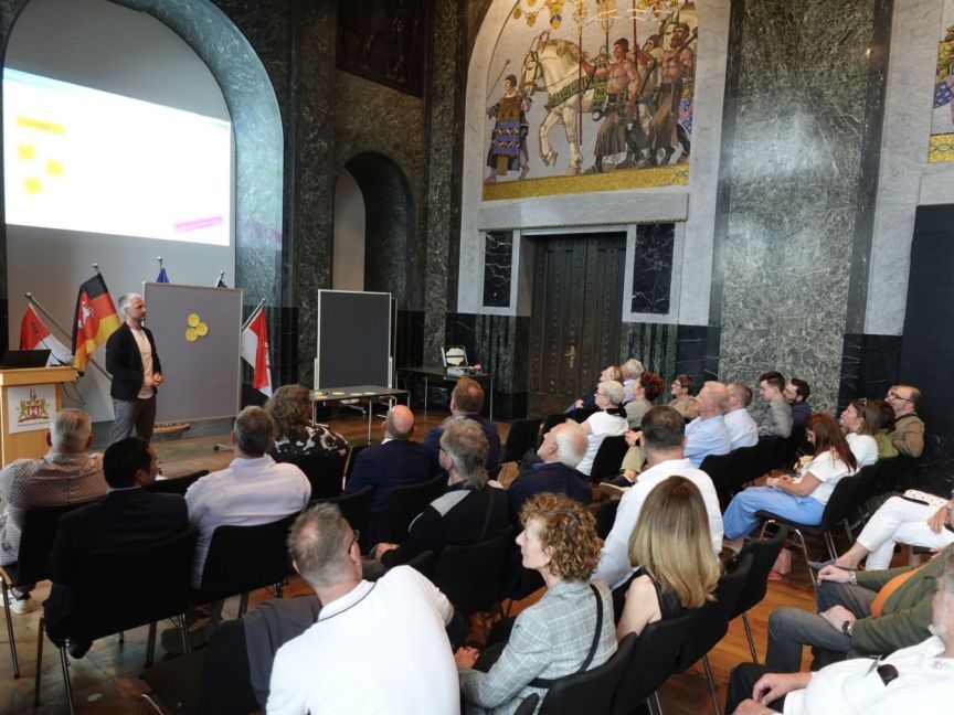 Ein Mann präsentiert vor Publikum in einem Saal mit Wandgemälden und Flaggen, dahinter eine Leinwand mit Folien.