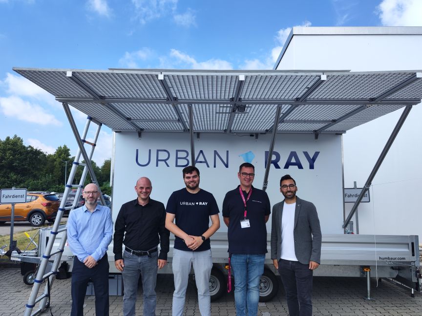 Fünf Männer stehen vor einem weißen Anhänger mit Solarpanelen und dem Logo „Urban Ray" bei bewölktem Himmel.