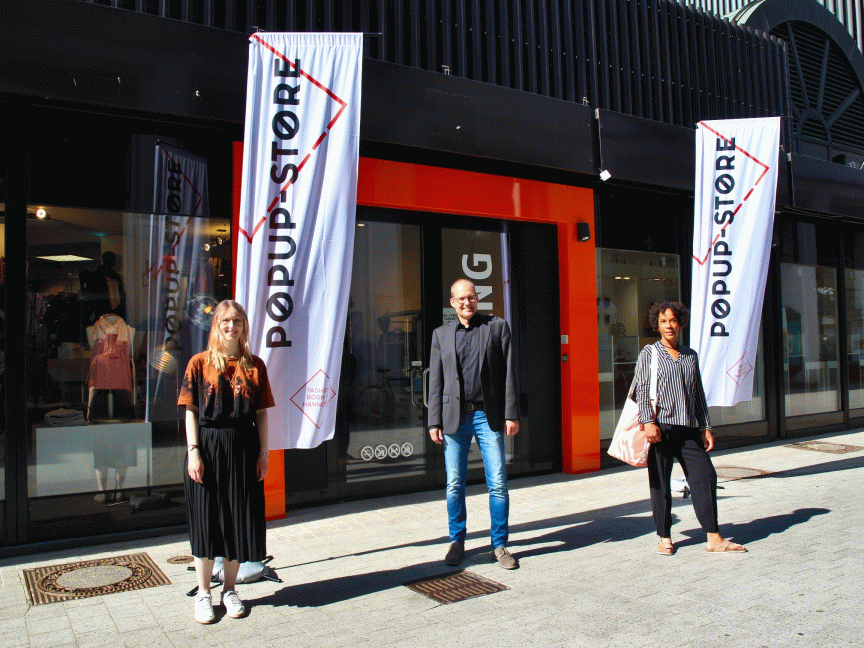 Drei Personen stehen vor einem Popup-Store mit weißen Fahnen und orangefarbenem Eingang.