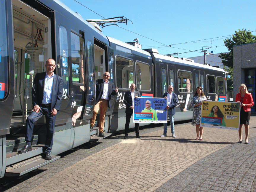 Gruppe von sieben Personen steht vor einer modernen Straßenbahn und hält Werbeplakate mit der Aufschrift „Digga muss los".