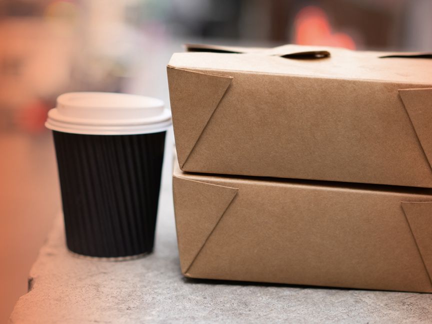 Zwei gestapelte Pappboxen und ein schwarzer Kaffeebecher mit weißem Deckel auf einer grauen Fläche.