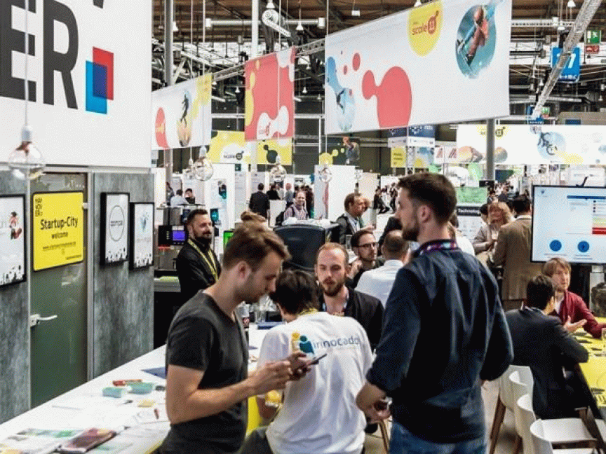 Messestand mit Besuchern in großer Halle, Schilder mit "Startup City" und bunten Logos, Personen stehen und sitzen an Tischen.