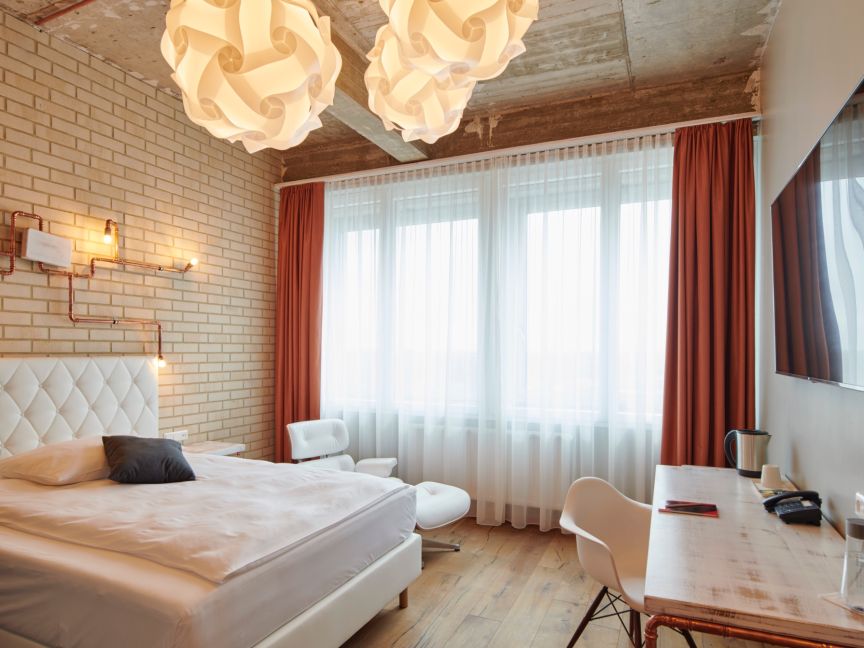 Hotelzimmer mit weißem Doppelbett, Ziegelwand, roten Vorhängen, Schreibtisch, TV und zwei Pendelleuchten.
